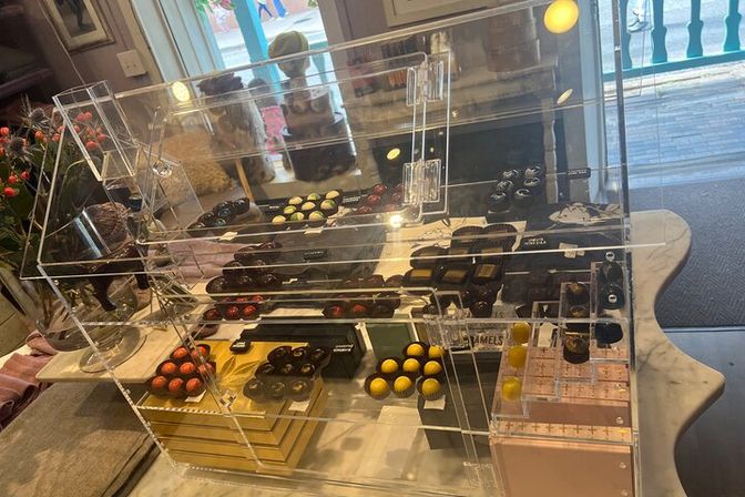 Acrylic display case of colorful artisanal chocolate bonbons and truffles arranged on black trays on a marble countertop by a sunlit shop window with a glimpse of a wooden boardwalk outside.