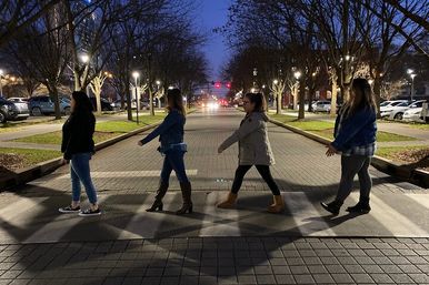 Four people playfully walking single-file across a lit pedestrian crosswalk on a tree-lined downtown street at dusk, with streetlights and parked cars in the background.