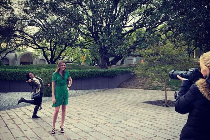 Photographer snaps a woman in a green dress posing in a tree-lined city plaza while a friend photobombs with a playful kick.