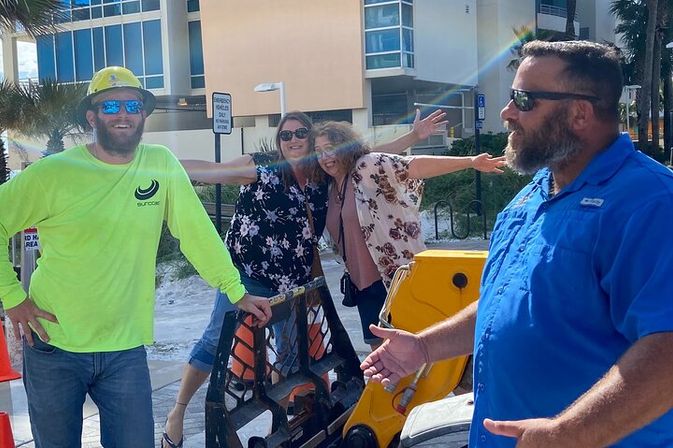 Smiling beachfront sidewalk crew in hi-vis gear and hard hat chatting near yellow equipment while two women pose with outstretched arms by palm trees and condos