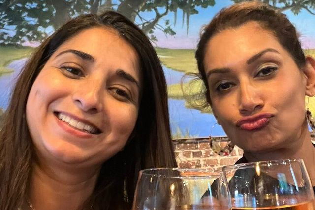 Two women taking a selfie in a casual restaurant, smiling and playfully posing with wine glasses in front of a painted landscape mural and exposed brick wall.