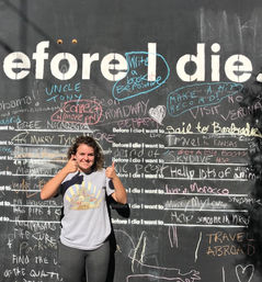 Smiling person giving two thumbs up in front of a giant 'Before I die' community chalkboard wall covered in colorful chalk messages and travel wishes — outdoor interactive public art