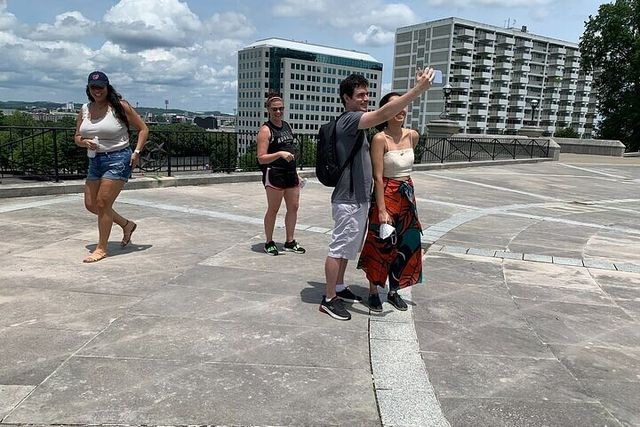 Group of four young adults on a sunny urban plaza; a man takes a selfie with a woman while two others stroll nearby, with downtown buildings and a partly cloudy sky in the background.