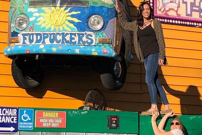 Colorful painted vintage VW van front mounted on a bright orange facade with sun mural; two women pose—one standing on a green ledge while the other supports her—playful outdoor attraction photo.