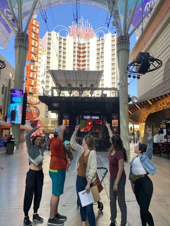 Cheerful group of five pointing up beneath a downtown Las Vegas light canopy toward a retro hotel tower and neon signage above a pedestrian stage
