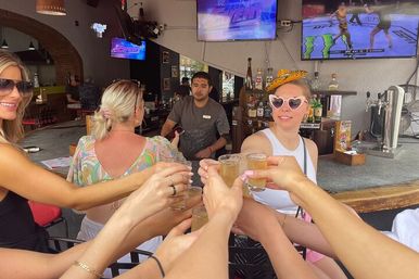 Friends clinking shot glasses at a sunny open-air bar counter, woman in heart-shaped sunglasses smiling, bartender and sports TVs in the background.