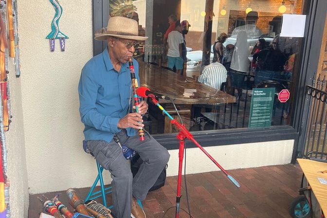 Street musician in a straw hat playing a colorful wooden flute into a microphone while seated on the sidewalk outside a busy café with patrons visible through the window.