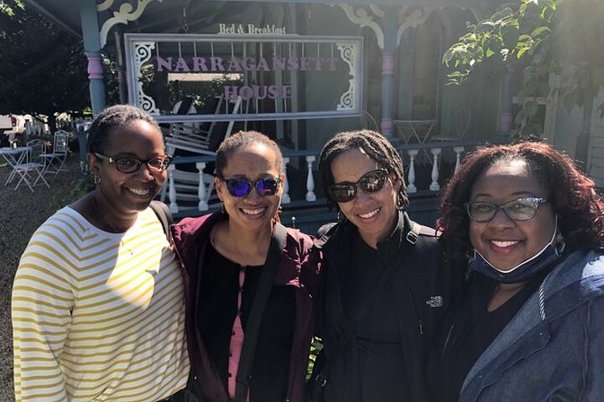Four friends smiling for a selfie in front of a pastel Victorian bed-and-breakfast porch with decorative trim and outdoor seating.