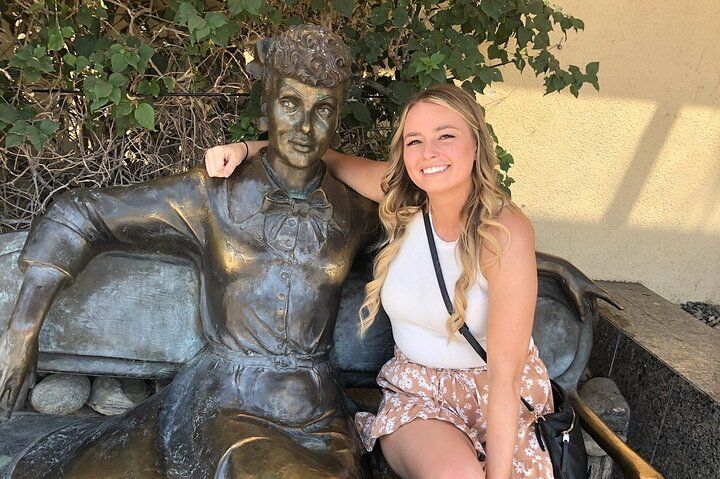 Smiling woman with long blonde hair sits on a bench with her arm around a seated bronze statue, ivy-covered backdrop and sunlit stucco wall.