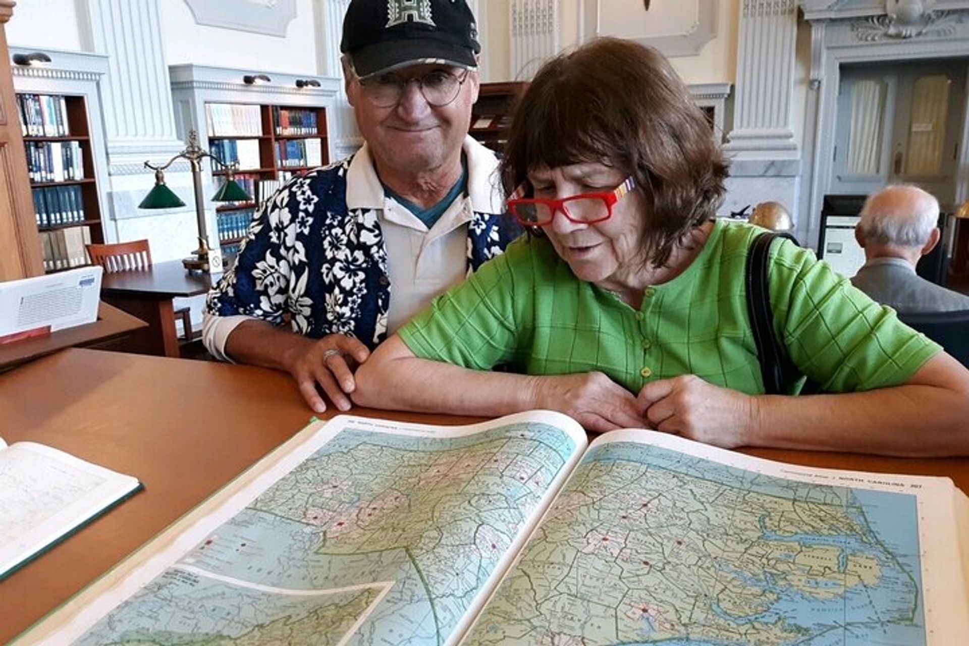 Two older adults leaning over an open atlas on a wooden library table, studying a large folded map with bookshelves and green reading lamps in the bright reading room — travel-planning vibe.