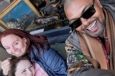 Three friends smiling for a close selfie outside an urban storefront window displaying framed artwork and small sculptures, wearing jackets and scarves.