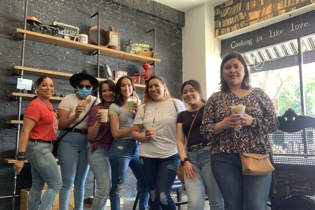 Seven friends smiling in a cozy urban café, standing by a brick wall with wooden shelves, each holding iced or hot coffee drinks under window light and a visible 'Cooking is like love' sign.
