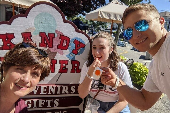 Three smiling people take a sunny outdoor selfie in front of a candy shop sign, one holding a fried-egg gummy candy while another wears blue mirrored sunglasses.
