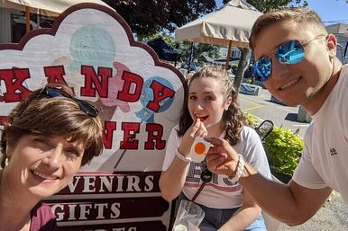 Group selfie of three people on a sunny small-town street in front of a colorful candy and souvenir shop sign, one holding a fried-egg-shaped candy with patio umbrellas and storefronts in the background.