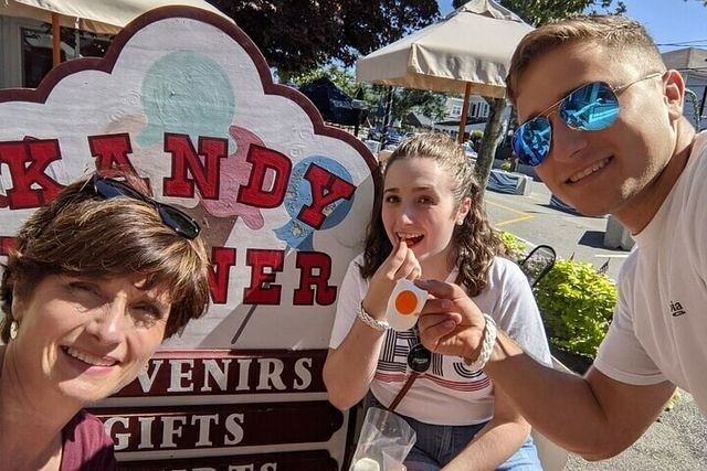 Group selfie of three people on a sunny small-town street in front of a colorful candy and souvenir shop sign, one holding a fried-egg-shaped candy with patio umbrellas and storefronts in the background.