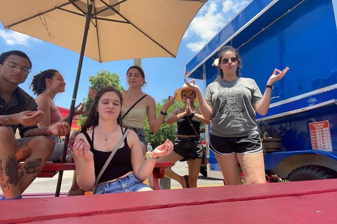 Group of six friends striking playful meditation poses under a patio umbrella at red picnic tables beside a blue food truck on a sunny summer day.