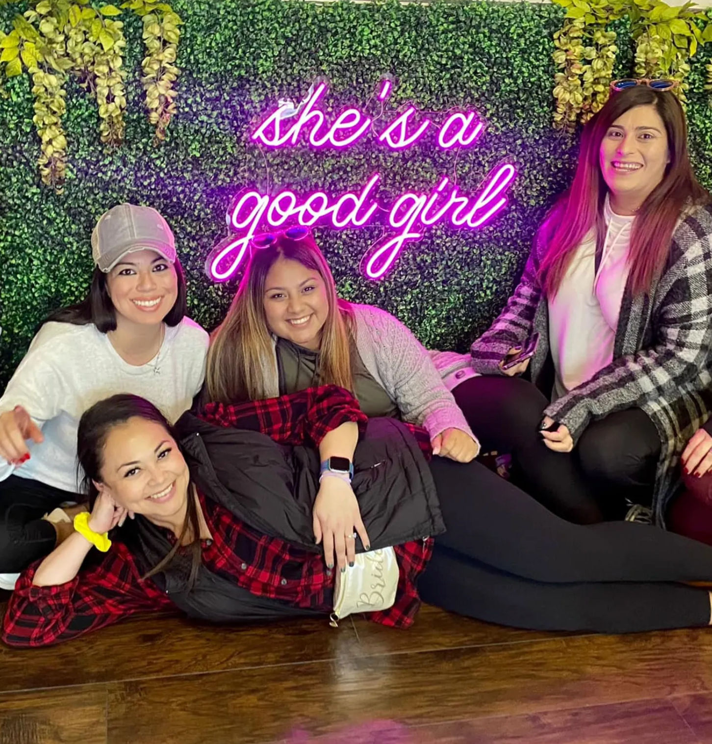 Four smiling women posing on a wooden floor in front of a lush green foliage wall with a bright pink neon sign reading 'she's a good girl' — casual outfits at an Instagrammable indoor photo backdrop.