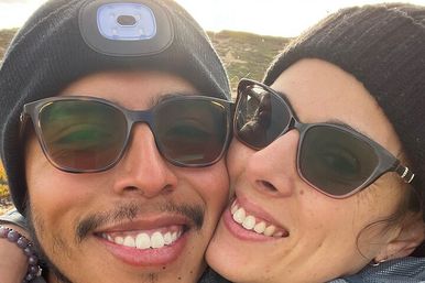 Close-up selfie of two smiling friends in sunglasses and knit beanies on a sunlit hillside, enjoying a nature hike.