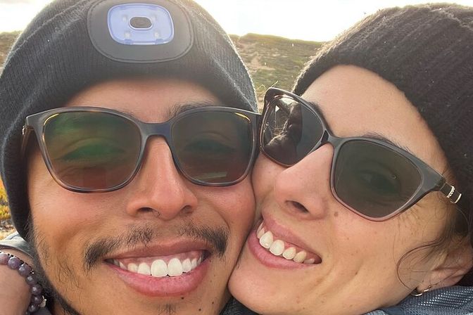 Close-up selfie of two smiling friends in sunglasses and knit beanies on a sunlit hillside, enjoying a nature hike.
