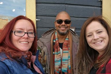 Three smiling people taking a selfie outside an urban storefront, wearing winter coats, a colorful scarf and sunglasses.