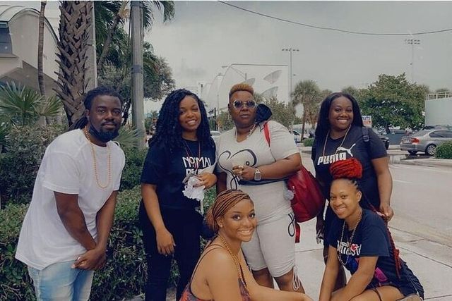 Six people smiling and posing on a palm-lined sidewalk outdoors, dressed in casual athletic wear and backpacks with parked cars and stadium lights in the background.