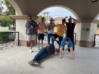 Seven friends striking playful poses—one lying down, one doing a plank—under an arched outdoor pavilion with palm trees and rocky hills in the background.