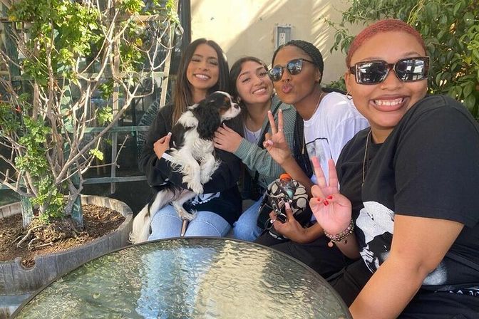 Four friends smiling on a sunny outdoor patio cafe, one holding a small black-and-white dog, two flashing peace signs, sunglasses and a round glass table with potted greenery in the background.