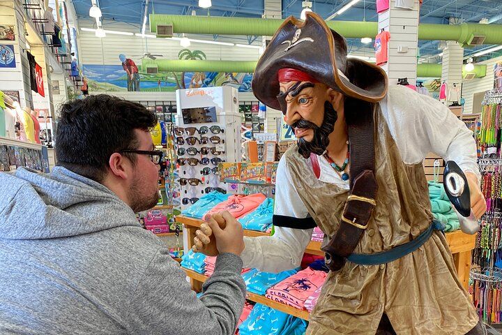 Person in a gray hoodie fist-bumping a life-size pirate statue inside a colorful coastal souvenir shop with sunglasses, beach t-shirts and accessories on display.