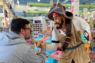 Person in a gray hoodie fist-bumping a life-size pirate statue inside a colorful coastal souvenir shop with sunglasses, beach t-shirts and accessories on display.