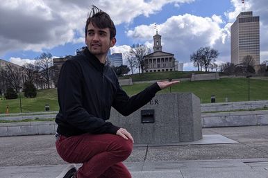 Young man crouching on an urban plaza, playfully pretending to hold a historic state capitol building on a grassy hill with downtown skyline and cloudy sky.