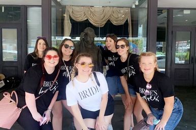 Seven women in matching bridal party shirts—bride in white and bridesmaids in black—smiling in sunglasses and posing for a bachelorette photo outside a theater-style entrance with glass doors and a statue behind them.