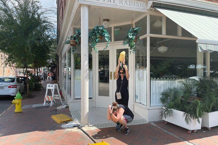 Playful street scene on a sunny downtown brick sidewalk outside a corner storefront with hanging plants and white columns: one person crouches covering their head while another stands behind holding a large ear of corn overhead; yellow fire hydrant and potted palms nearby.
