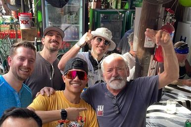 Group of friends at an outdoor bar patio, daytime — older bearded man raising his arm and smiling, younger man in a yellow shirt and mirrored sunglasses pointing, three companions in hats behind, cooler and bar gear in the background.