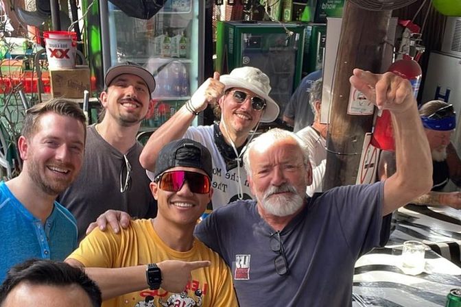 Group of friends at an outdoor bar patio, daytime — older bearded man raising his arm and smiling, younger man in a yellow shirt and mirrored sunglasses pointing, three companions in hats behind, cooler and bar gear in the background.