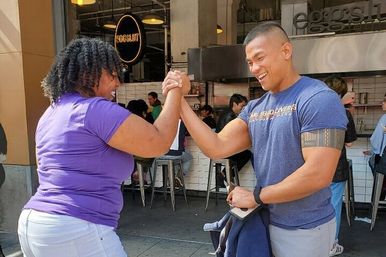 Playful arm-wrestling handshake between two people laughing outside a busy sidewalk café — woman in a purple shirt and man in a blue shirt with a tattoo.