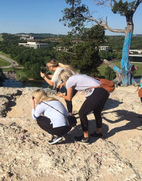 Three visitors crouch to snap photos at a rocky hilltop overlook, bright blue-painted tree framing a river and green valley under a clear blue sky