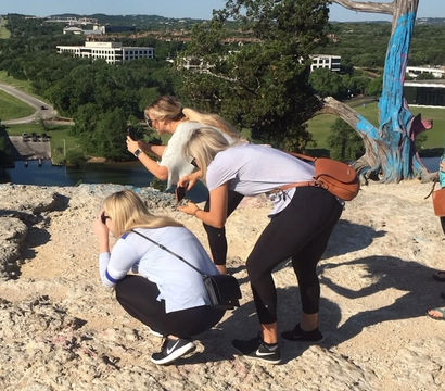Three visitors crouch to snap photos at a rocky hilltop overlook, bright blue-painted tree framing a river and green valley under a clear blue sky