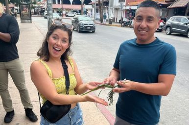 Smiling woman in a yellow top and a man in a blue shirt exchange a sprig of green plant on a busy street with shops and parked cars