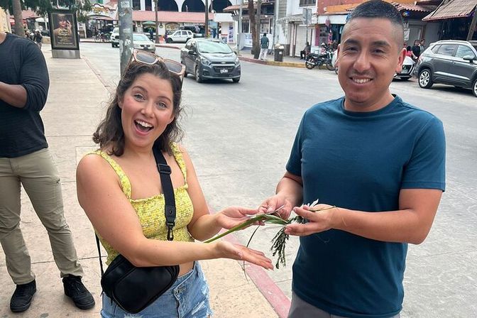 Smiling woman in a yellow top and a man in a blue shirt exchange a sprig of green plant on a busy street with shops and parked cars