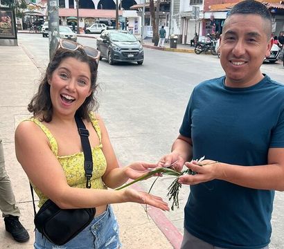 Smiling woman in a yellow top and a man in a blue shirt exchange a sprig of green plant on a busy street with shops and parked cars