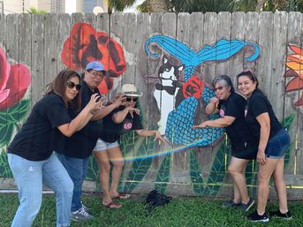 Five adults in black shirts pose playfully like claws in front of a colorful wooden fence mural of a cat-mermaid and oversized flowers on a sunny day with palm trees in the background.