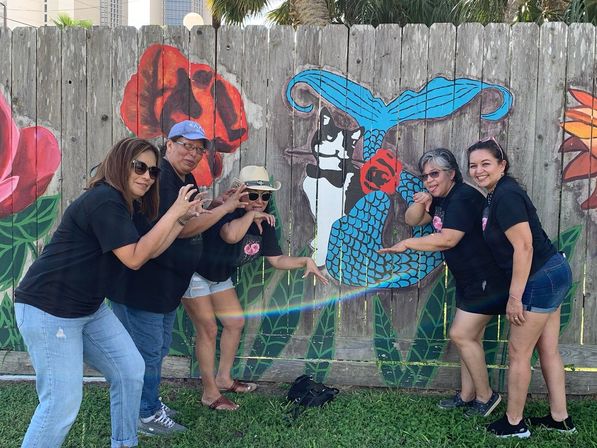 Five adults in black shirts pose playfully like claws in front of a colorful wooden fence mural of a cat-mermaid and oversized flowers on a sunny day with palm trees in the background.