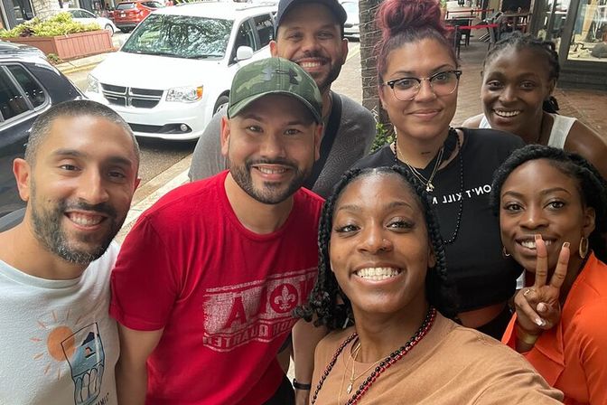 Seven smiling friends take a cheerful selfie on a lively city sidewalk outside storefronts, wearing casual summer outfits and one person flashing a peace sign.