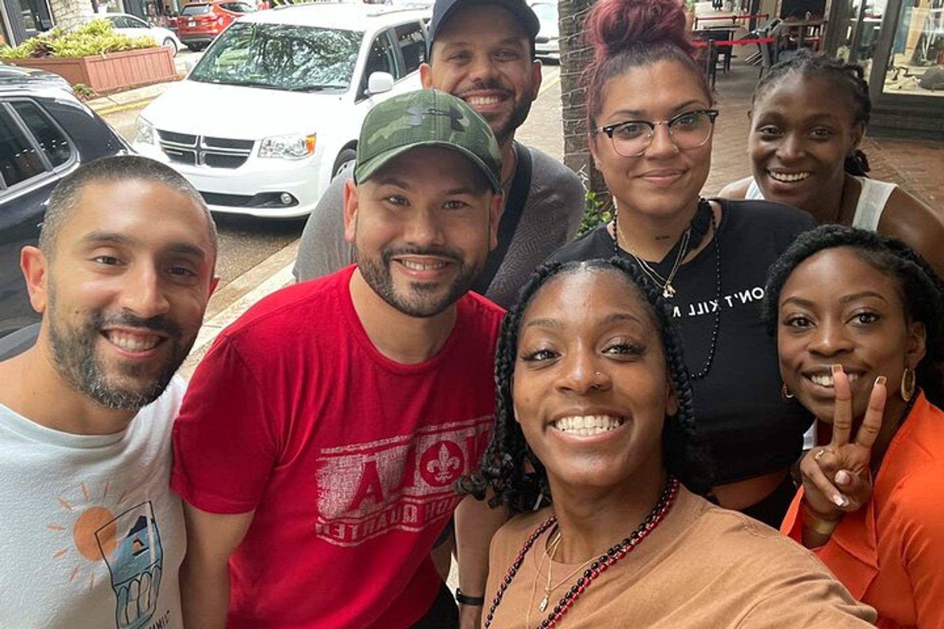 Seven smiling friends take a cheerful selfie on a lively city sidewalk outside storefronts, wearing casual summer outfits and one person flashing a peace sign.
