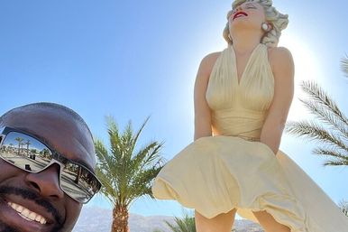 Person smiling in sunglasses taking a selfie beside a giant retro statue of a laughing woman in a halter dress with her skirt blown up, backlit by the sun with palm trees and mountains under a clear blue sky.