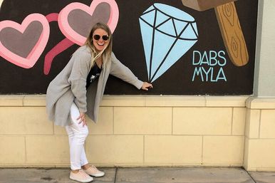 Woman in sunglasses and gray cardigan posing beside colorful street-art mural of pink hearts and a blue diamond on a downtown sidewalk wall