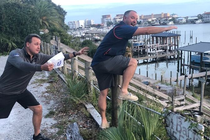 Two men at a coastal marina — one playfully reaching while the other climbs over a wooden railing toward docks and boat slips, with palm trees and waterfront condos in the background.
