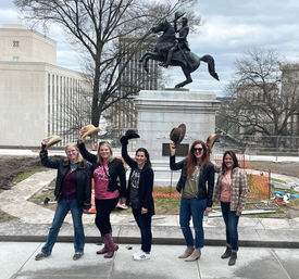 Five women smiling and lifting cowboy hats while posing on a city plaza sidewalk in front of an equestrian monument and bare winter trees on an overcast day.