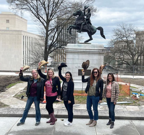 Five women smiling and lifting cowboy hats while posing on a city plaza sidewalk in front of an equestrian monument and bare winter trees on an overcast day.