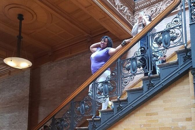 Smiling person in a purple top posing on an ornate wrought-iron staircase with wooden handrail inside a grand historic interior — wood-paneled ceiling, brick walls and a hanging pendant light.